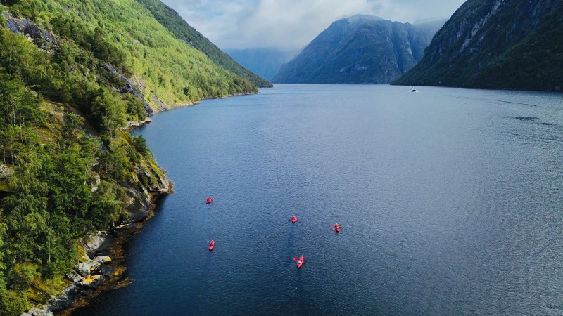 Kayakers in red on a tranquil fjord surrounded by green mountains under a cloudy blue sky.
