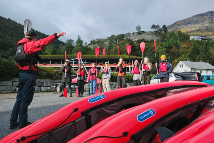 Instructor and group hold paddles near red kayaks with forested hillside in background.