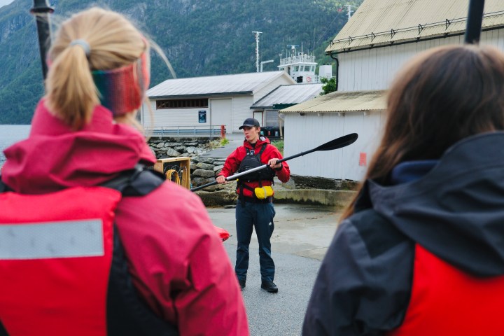 Instructor in red jacket demonstrates with paddle to two people by water and buildings.