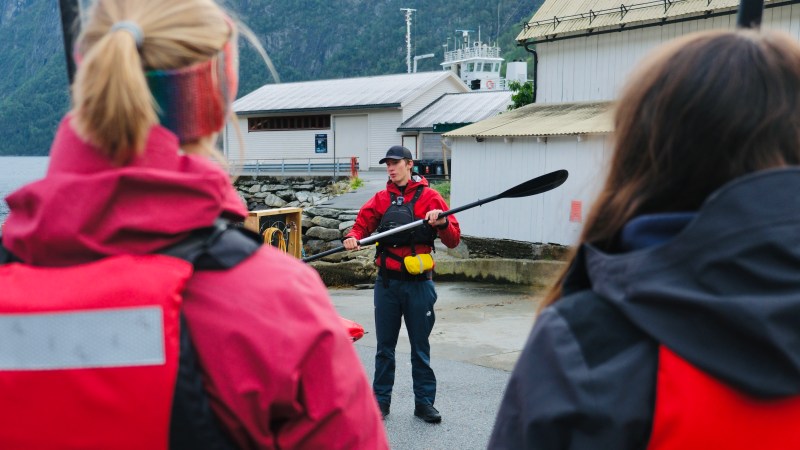 Instructor in red jacket demonstrates with paddle to two people by water and buildings.