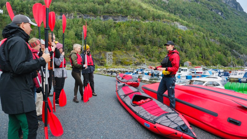 Group receives kayak instruction near colorful boats and forested hills.