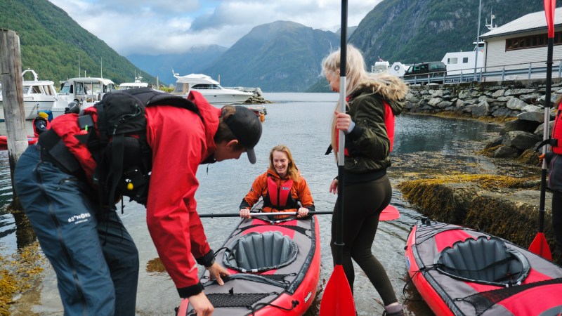 Three people preparing kayaks by a fjord with mountains and boats in the background.