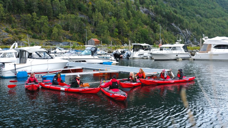 Group of kayakers in red kayaks near docked boats with forested hill in background.