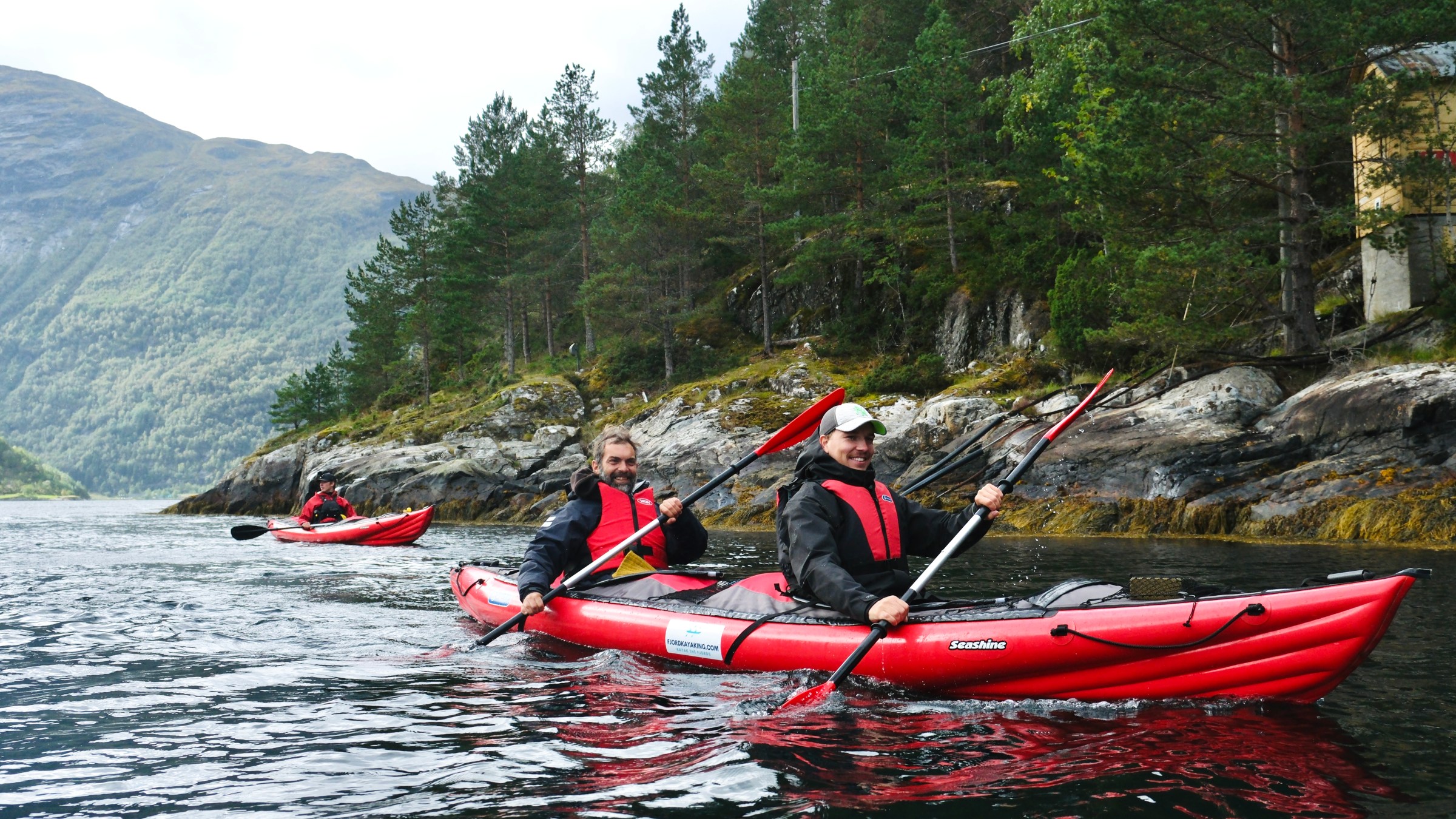 Two people kayaking in red kayaks on a scenic fjord with pine trees and mountains in the background.