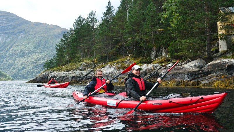 Two people kayaking in red kayaks on a scenic fjord with pine trees and mountains in the background.