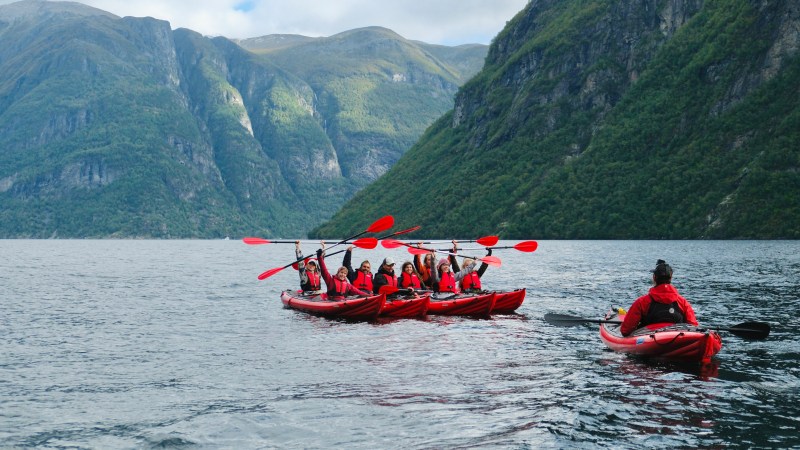 Group kayaking in red kayaks on a lake with green mountain backdrop.
