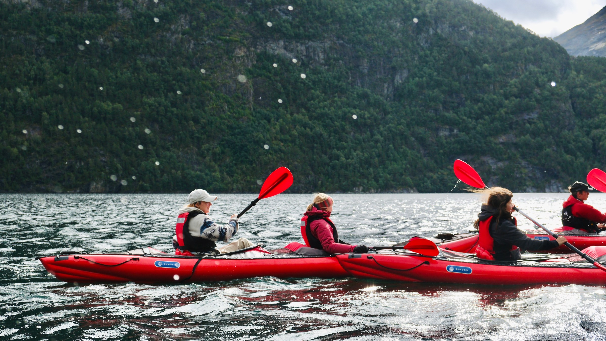 People kayaking in red kayaks on a lake, surrounded by forested hills.