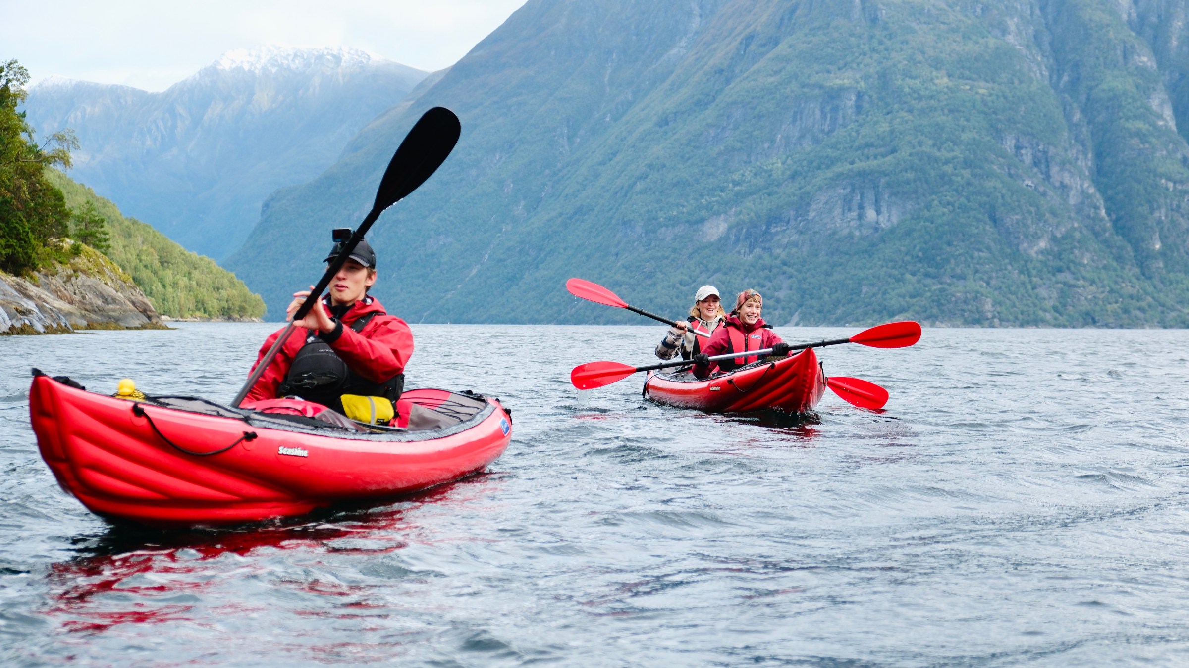 Three people kayaking in red kayaks on a lake surrounded by mountains.