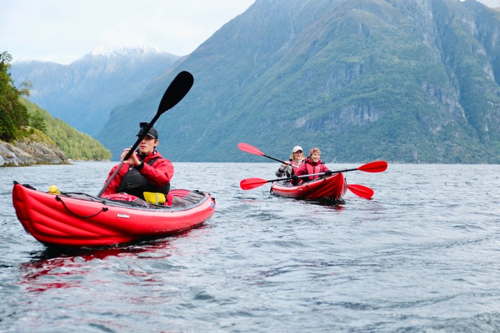Three people kayaking in red kayaks on a lake surrounded by mountains.