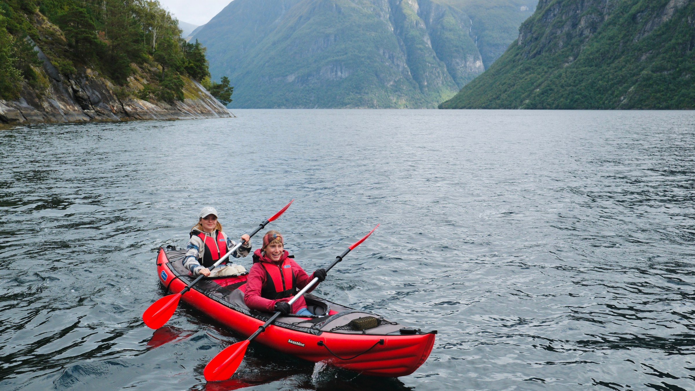 Two people in a red kayak paddle on a fjord with steep mountains in the background.