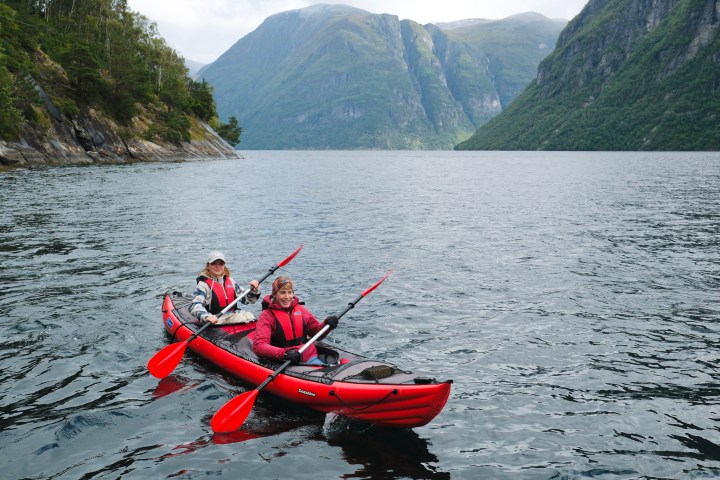 Two people in a red kayak paddle on a fjord with steep mountains in the background.