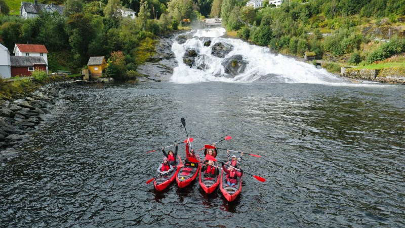Four kayakers with red paddles on a river near a waterfall and houses on a hillside.