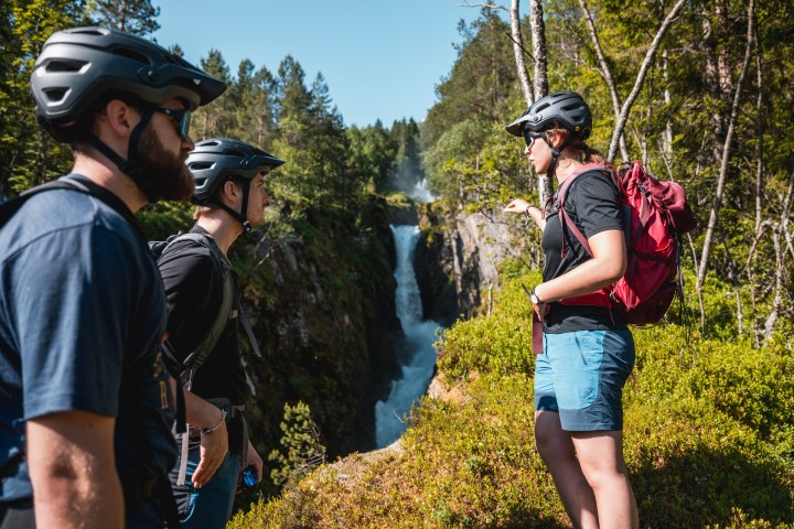 Three people with helmets view a waterfall in a forested area.