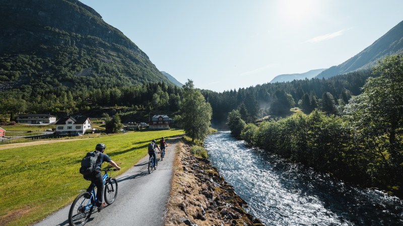 Cyclists on a rural path by a river and mountains under a clear blue sky.