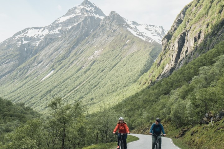Two cyclists ride on a scenic mountain road with snow-capped peaks in the background.