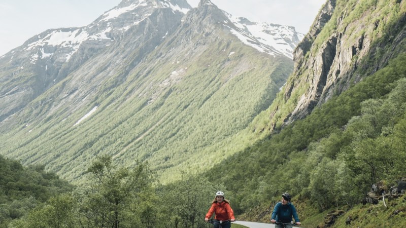 Two cyclists ride on a scenic mountain road with snow-capped peaks in the background.