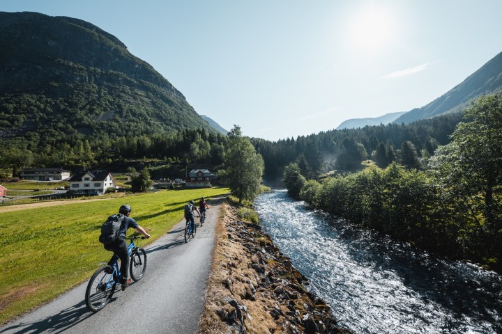 Cyclists ride along a riverside path with mountains and trees under a clear blue sky.