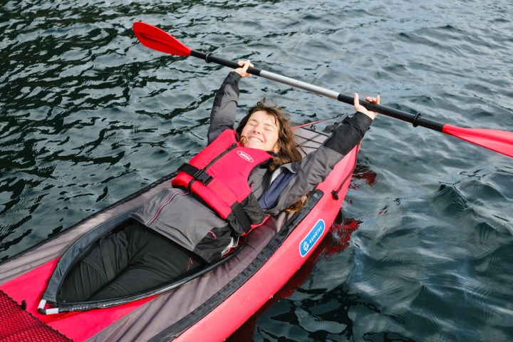 Smiling person relaxing in a red kayak, holding a paddle above their head on calm water.