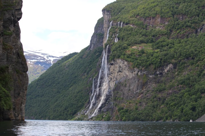 Tall waterfall cascading down a lush green cliff into a fjord under a partly cloudy sky.