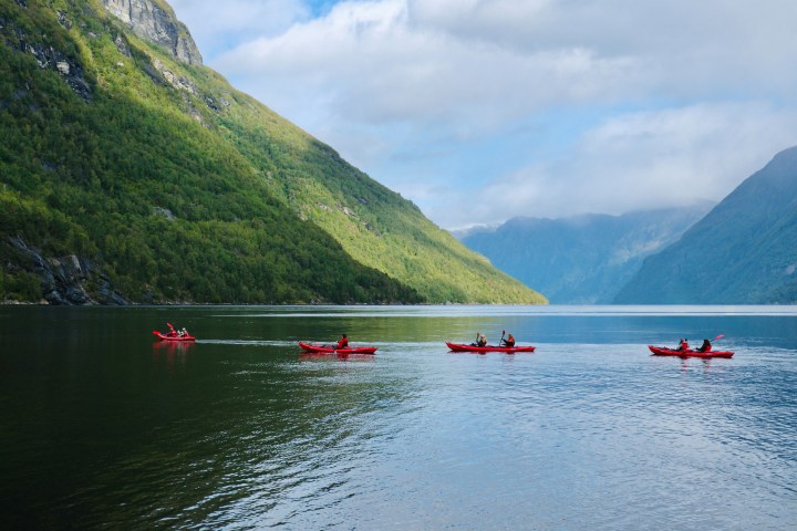 People kayaking in red boats on a serene fjord with lush green mountains.
