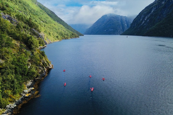 Red kayaks on a blue lake with green hills and distant mountains under a cloudy sky.