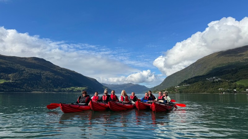 Group kayaking on calm lake with mountains and cloudy sky in background.