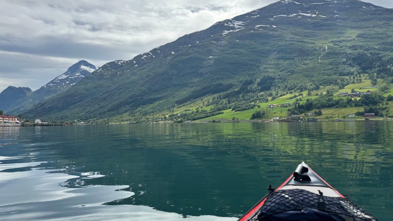 View from kayak on serene water, with mountains and scattered houses in background under cloudy sky.