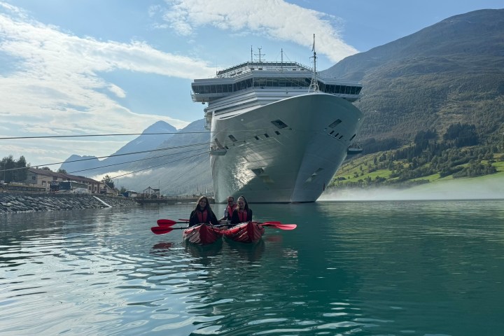 Two people kayaking in front of a large cruise ship on a calm lake with mountains in the background.