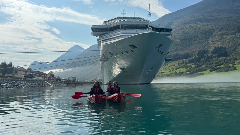 Two people kayaking in front of a large cruise ship on a calm lake with mountains in the background.