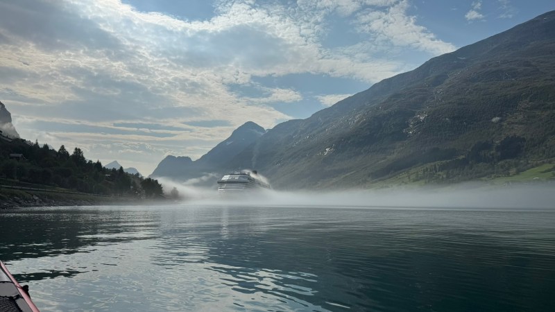 Cruise ship in misty fjord surrounded by mountains under cloudy sky.