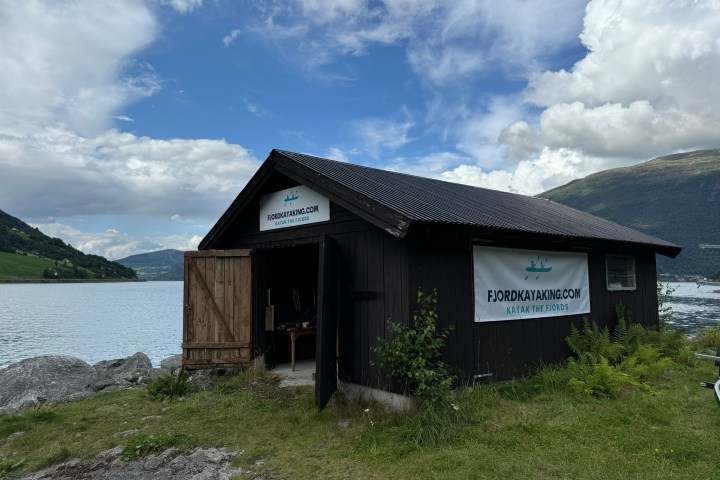 Wooden shed by fjord with sign 'Fjordkayaking.com'. Open door, grassy foreground, cloudy sky, scenic backdrop.