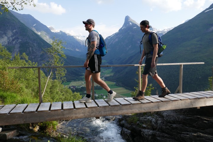 Two hikers on a wooden bridge with mountains in the background.