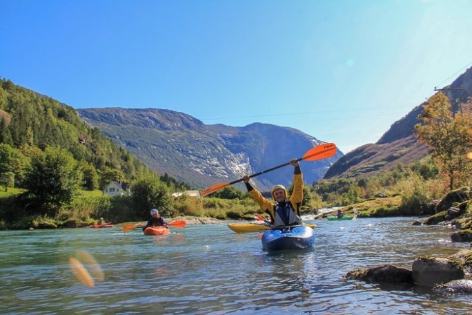 People kayaking on a river surrounded by mountains and trees under a clear blue sky.